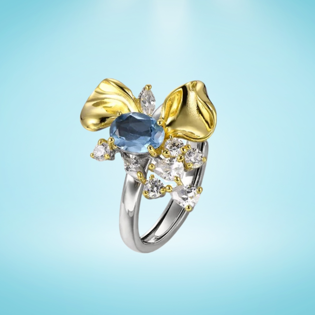 Two-tone floral ring with a blue oval gemstone, gold petal accents, and white crystal details on a silver band, set against a light blue background