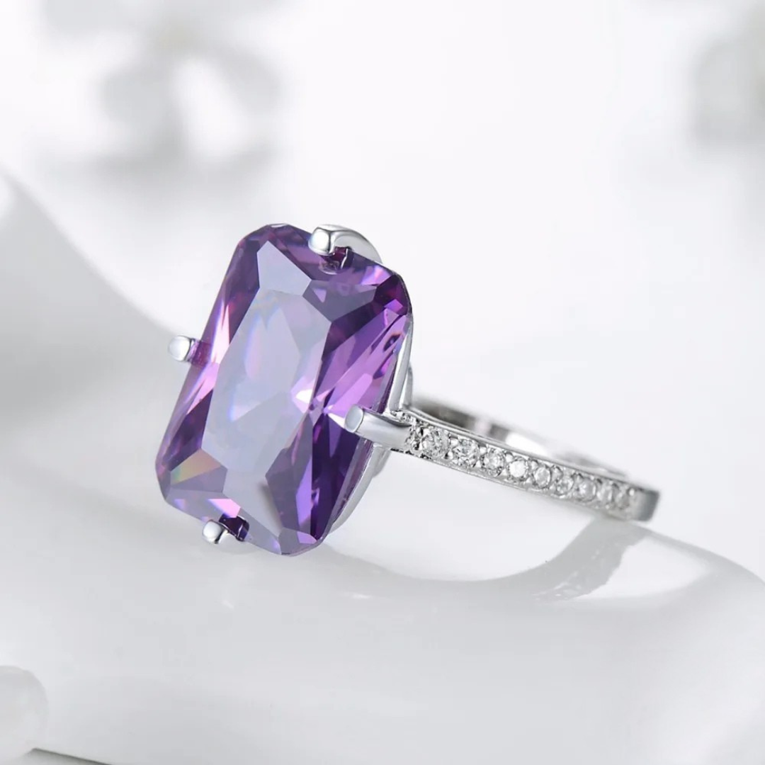 Sterling silver ring with a large rectangular-cut purple gemstone and pavé-set clear stones along the band, displayed on a light blue background