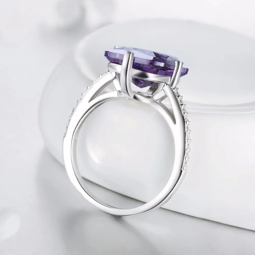 Sterling silver ring with a large rectangular-cut purple gemstone and pavé-set clear stones along the band, displayed on a light blue background