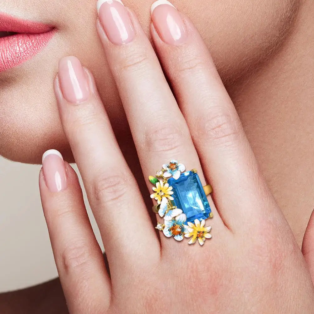 Gold ring with a large rectangular blue gemstone and colorful enamel flowers arranged around it