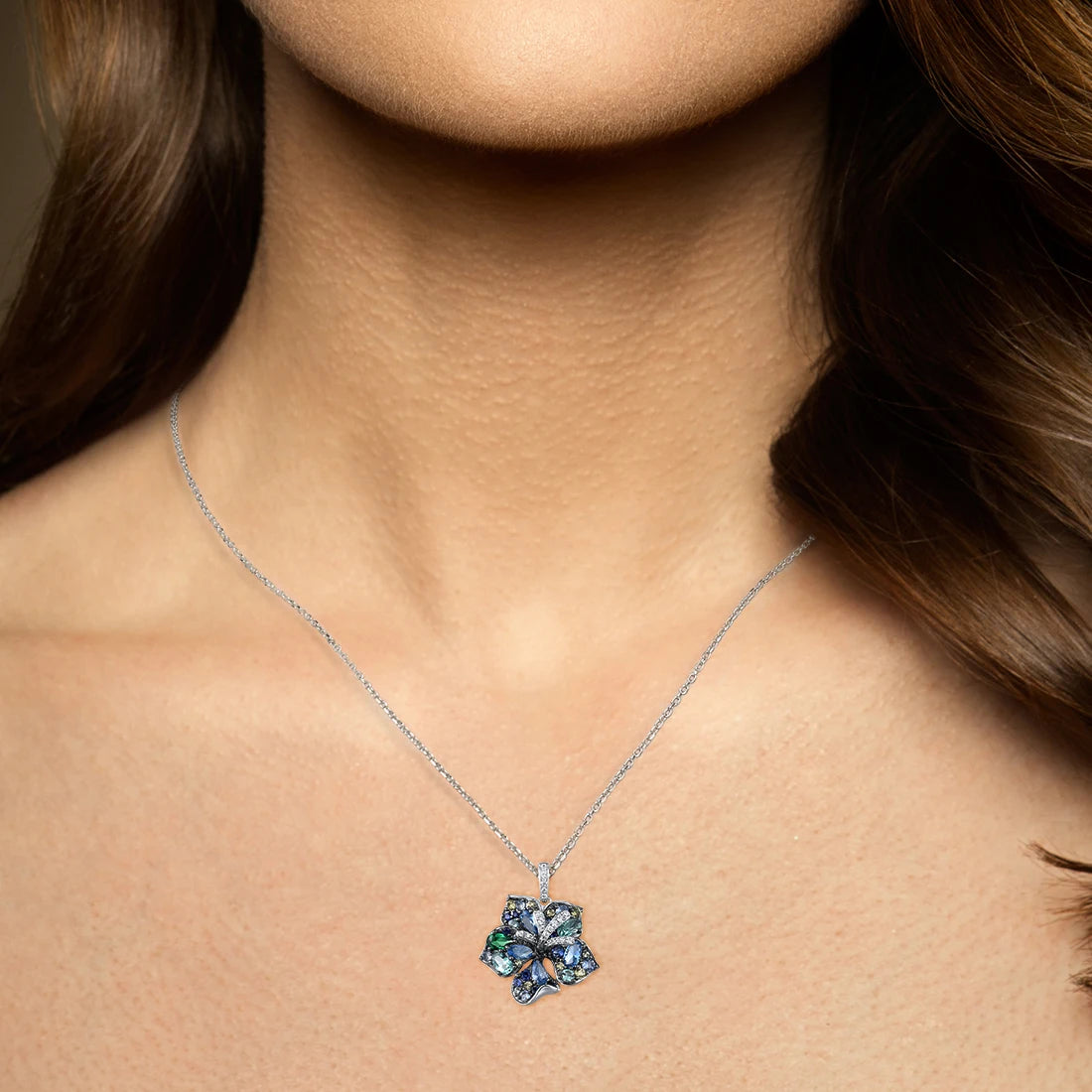 Silver pendant shaped like a stylised leaf or flower, set with a mix of blue, green, and yellow gemstones in varying cuts, with pavé crystal accents