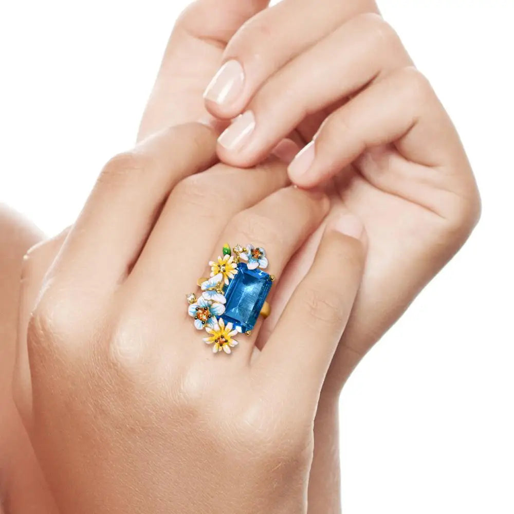 Gold ring with a large rectangular blue gemstone and colorful enamel flowers arranged around it