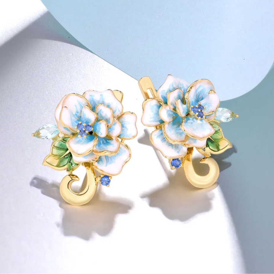 Gold-tone enamel earrings shaped like white and sky blue flowers with green leaves and sparkling stones.