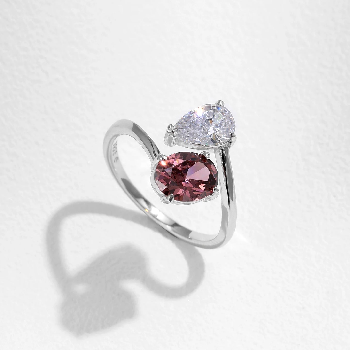 Silver ring with a split band featuring a clear pear-cut stone and a deep rose oval gem, shown on a red background from the Rossa collection.
