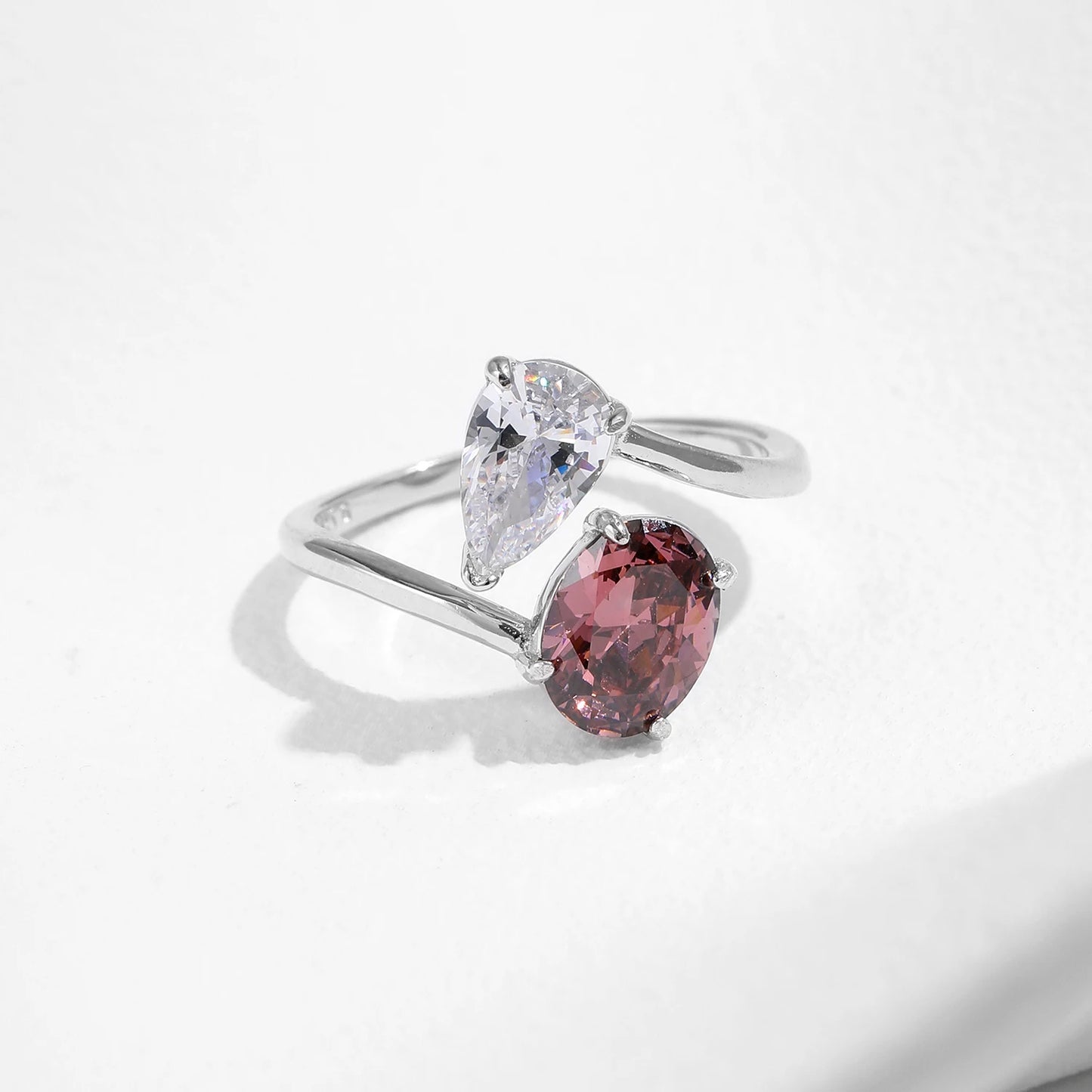 Silver ring with a split band featuring a clear pear-cut stone and a deep rose oval gem, shown on a red background from the Rossa collection.