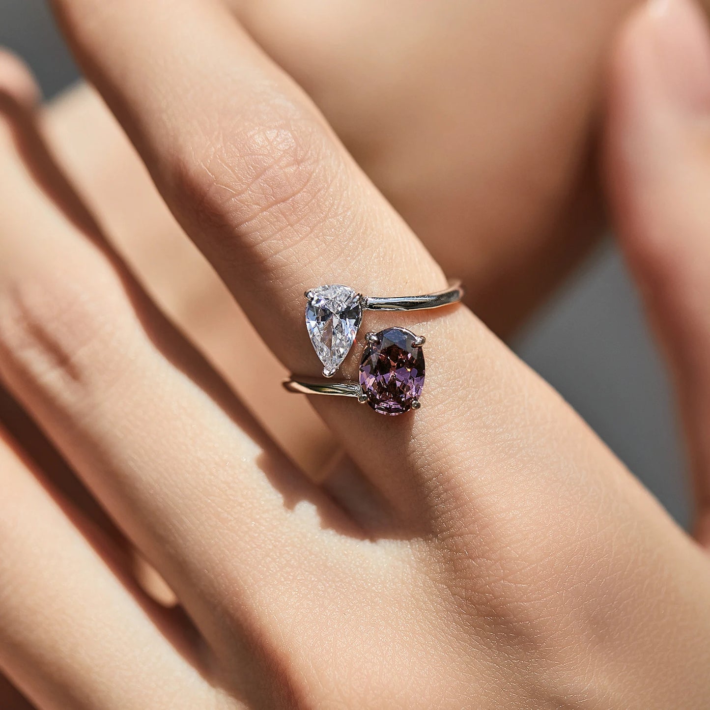 Silver ring with a split band featuring a clear pear-cut stone and a deep rose oval gem, shown on a red background from the Rossa collection.