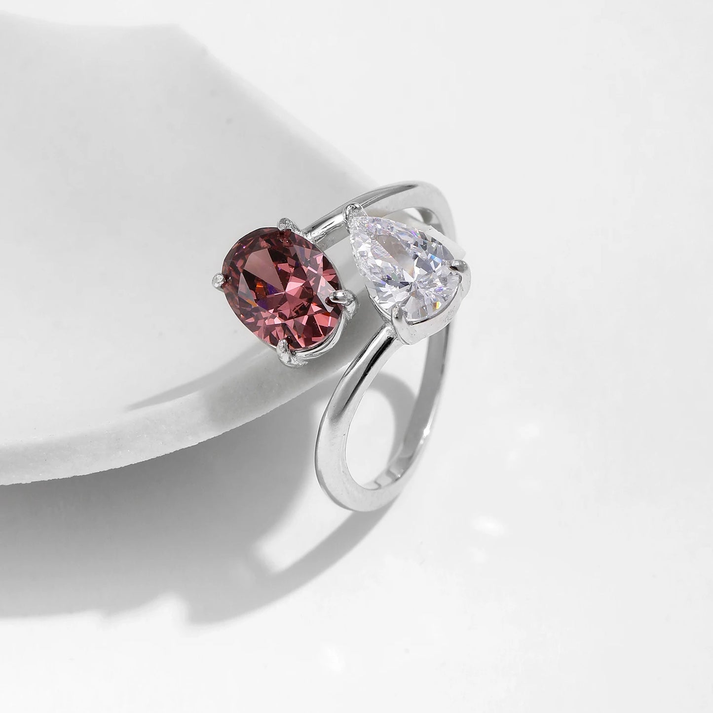 Silver ring with a split band featuring a clear pear-cut stone and a deep rose oval gem, shown on a red background from the Rossa collection.