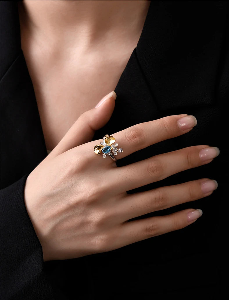Two-tone floral ring with a blue oval gemstone, gold petal accents, and white crystal details on a silver band, set against a light blue background