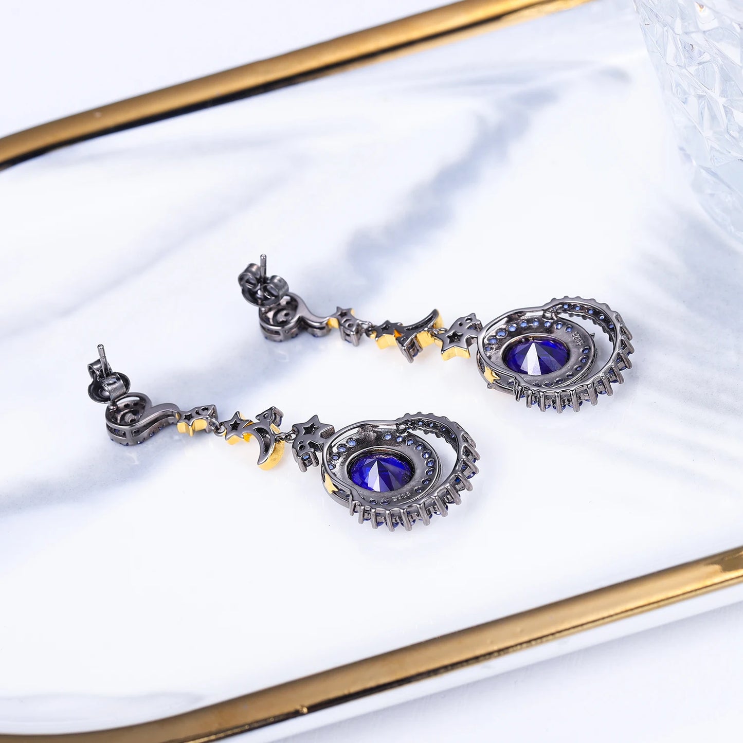 Dangling earrings with gold moon and star charms, deep blue oval stones, and pavé-set blue crystals in a celestial design