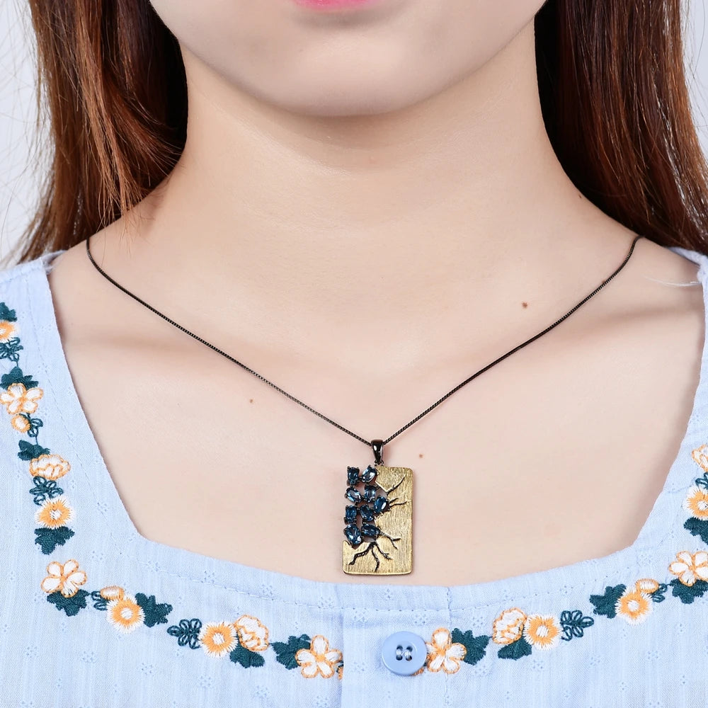 Rectangular gold pendant featuring dark branch-like details set with oval blue gemstones, hanging from a black chain