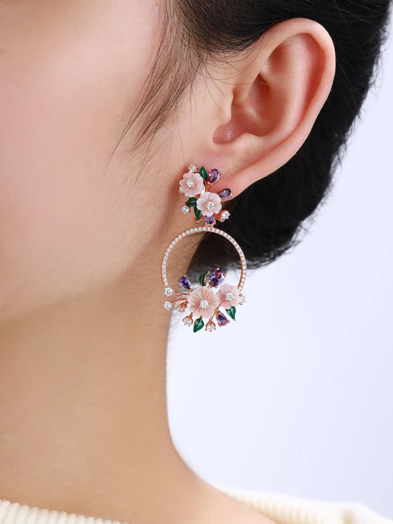 Infiorescenza hoop earrings featuring peach-toned flowers, purple and white stones, and green enamel leaves in an ornate floral arrangement.
