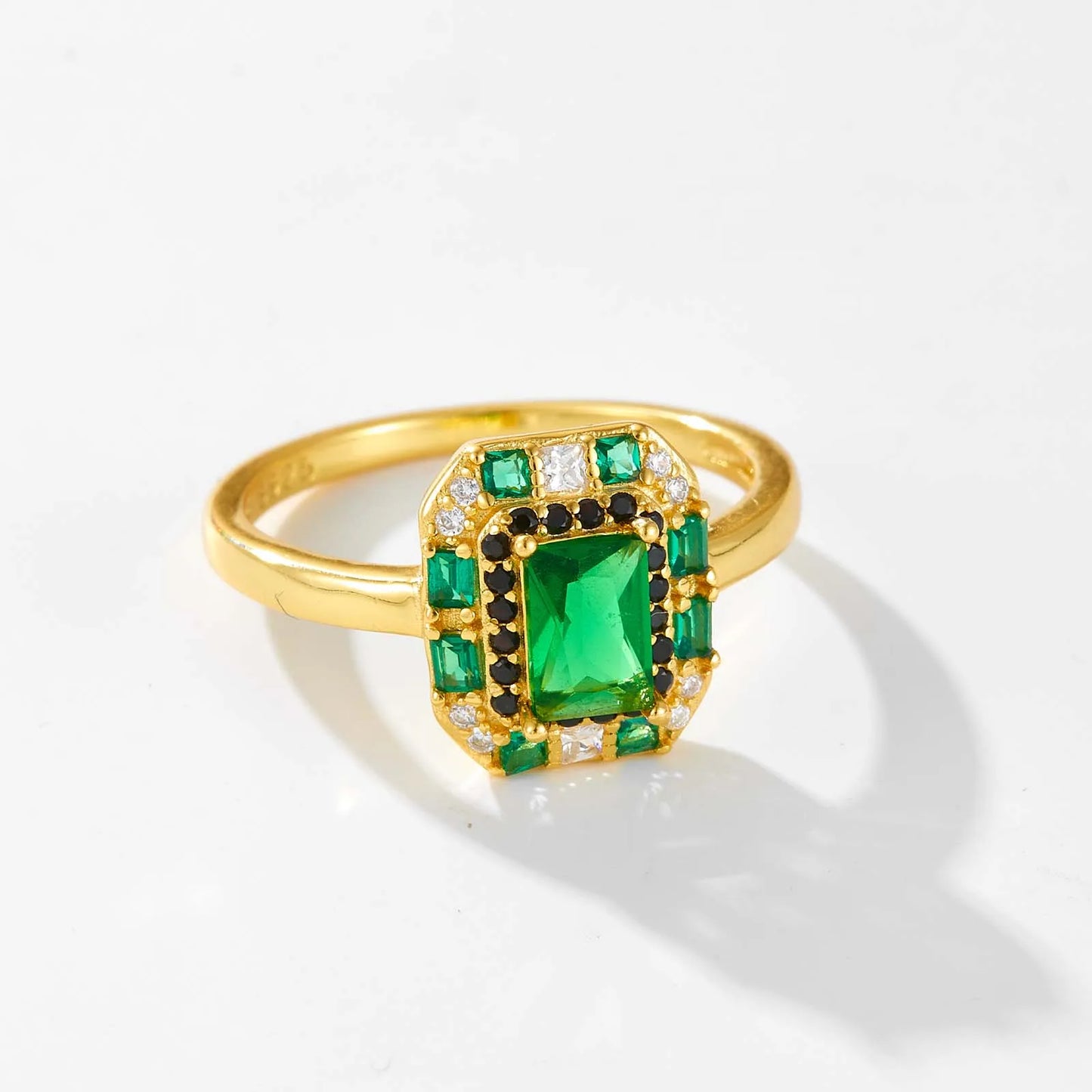 Gold ring with a central emerald-green rectangular stone, surrounded by alternating black and white crystals and square-cut green gems.