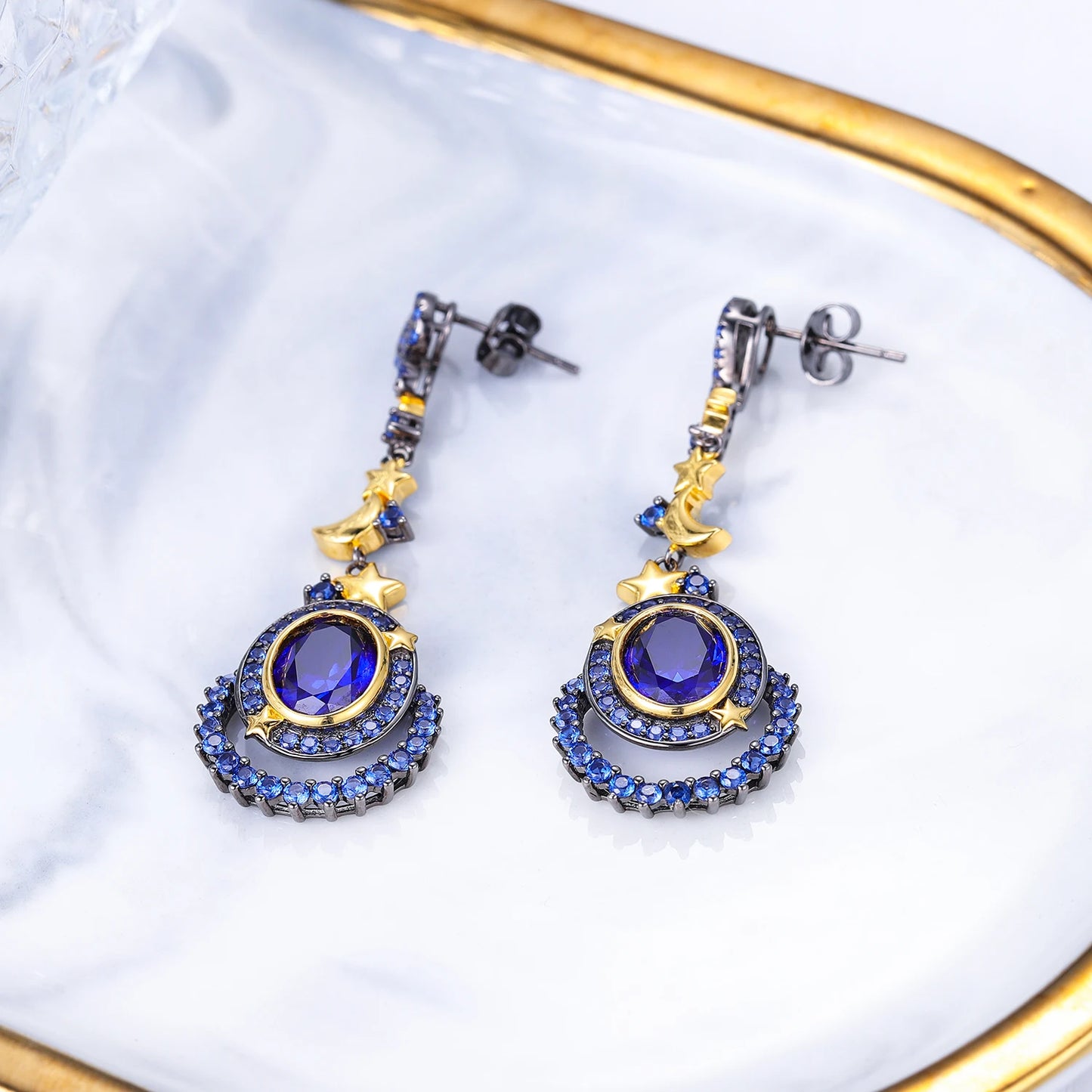 Dangling earrings with gold moon and star charms, deep blue oval stones, and pavé-set blue crystals in a celestial design