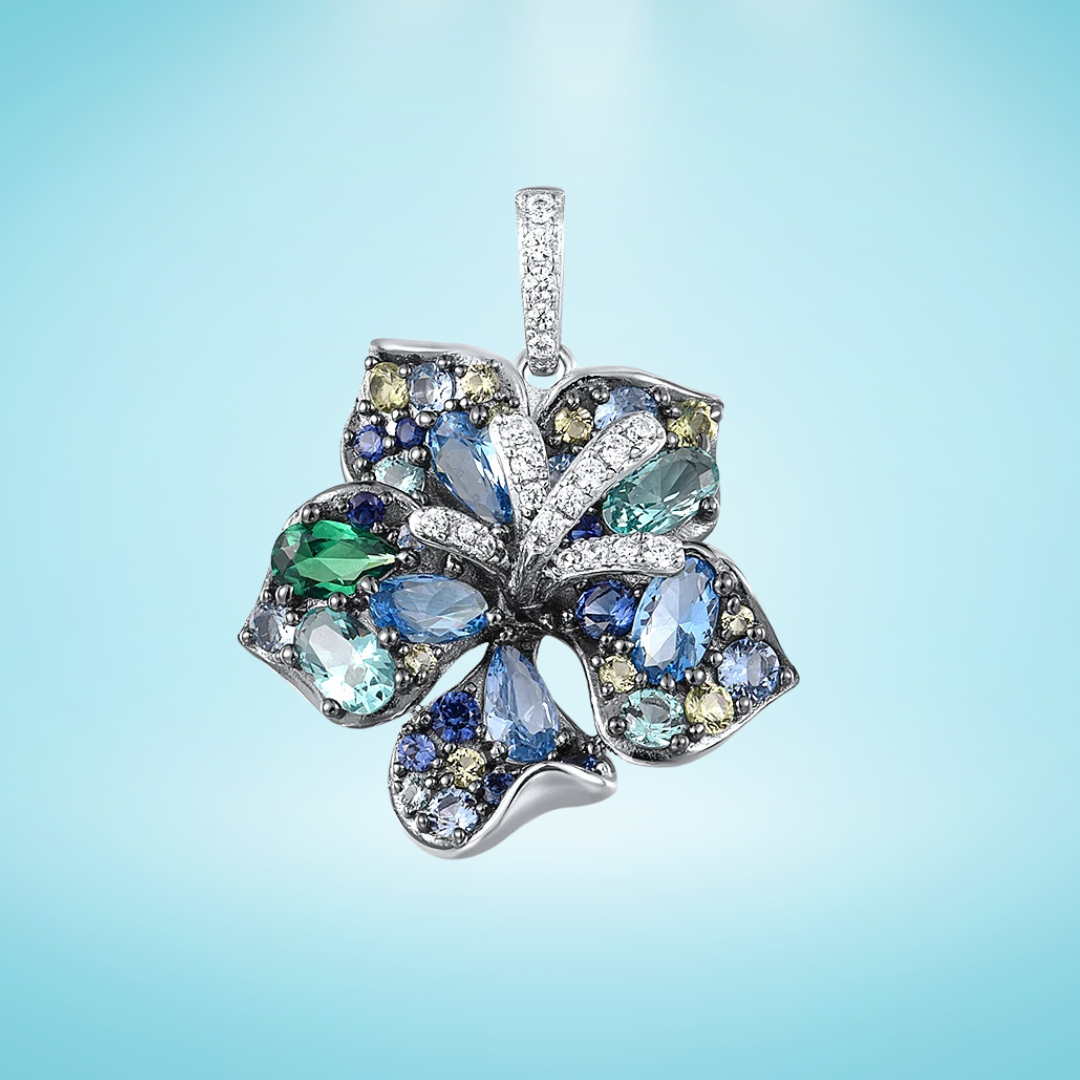 Silver pendant shaped like a stylised leaf or flower, set with a mix of blue, green, and yellow gemstones in varying cuts, with pavé crystal accents, on a light blue background