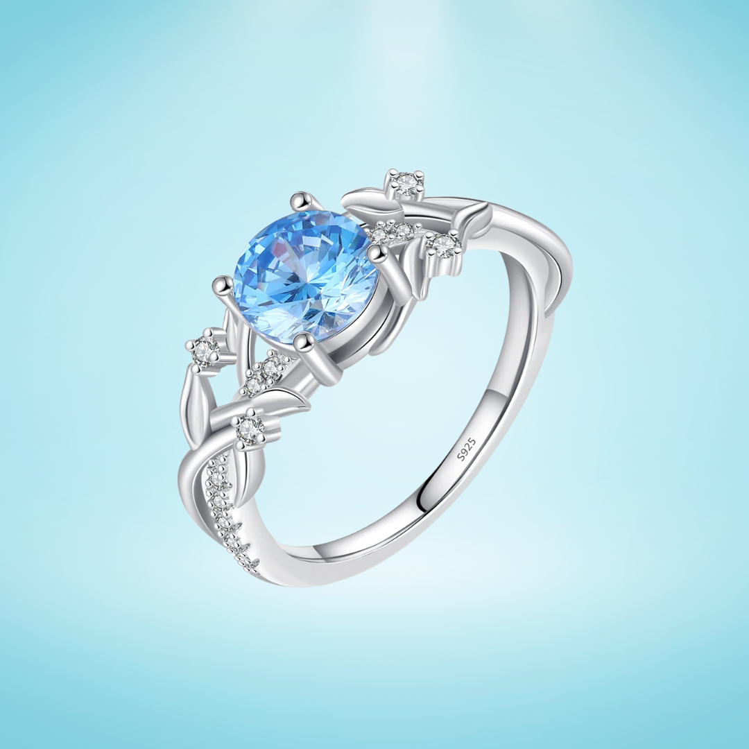 Sterling silver ring with a round sky blue gemstone and flowing leaf-like band details accented by small clear stones, set on a light blue background