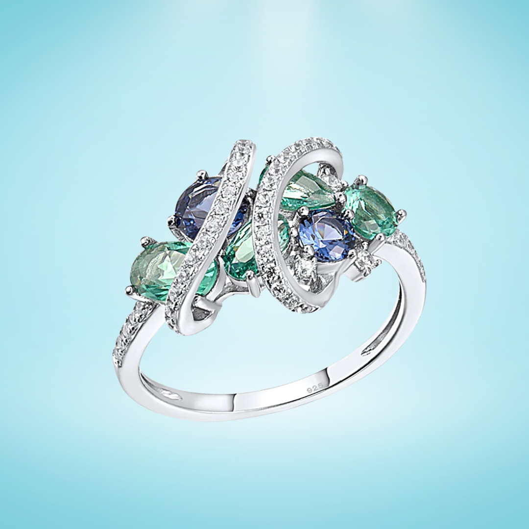 Sterling silver ring with swirling bands of pavé stones and clustered green and blue round-cut gemstones, displayed on a light blue background