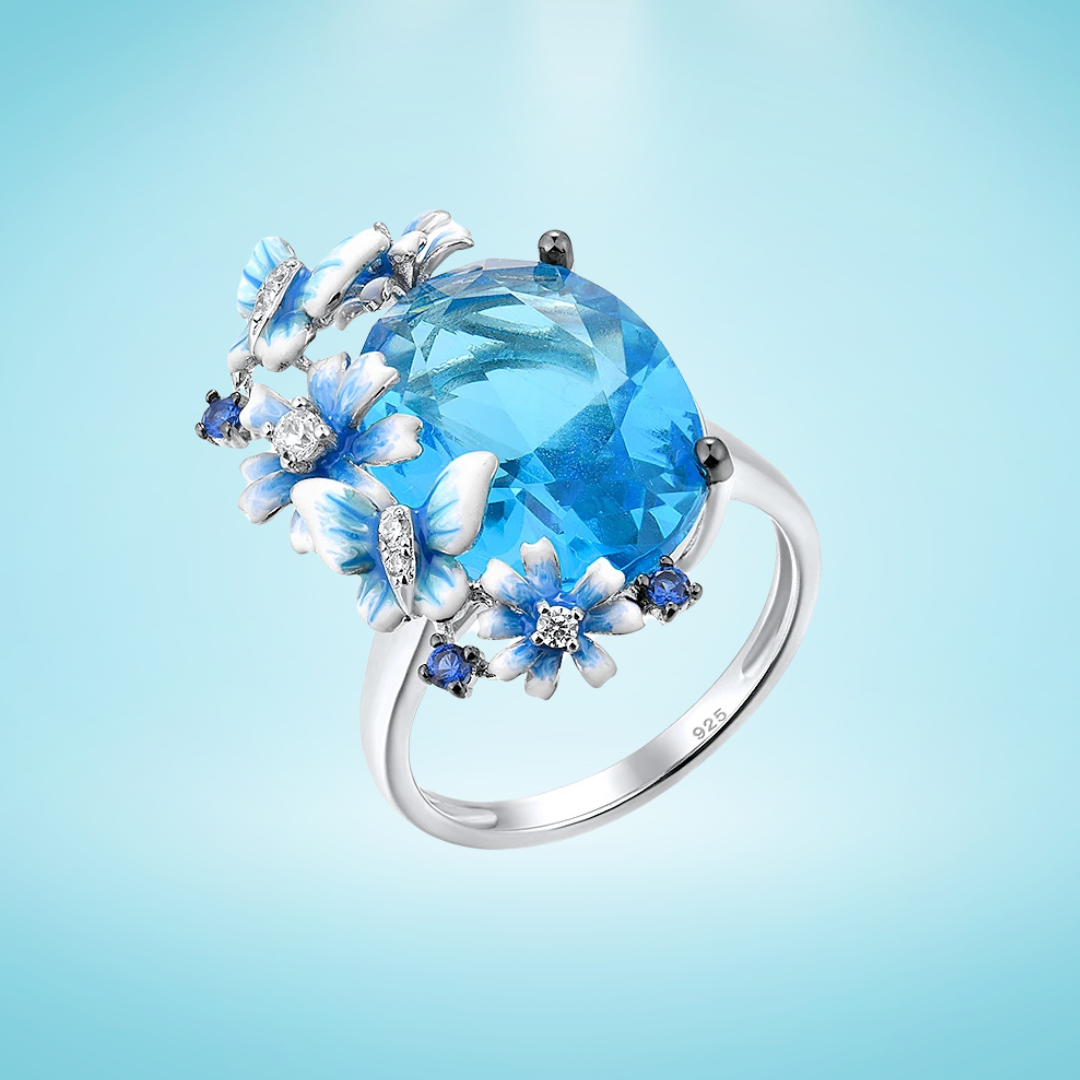 Silver ring with a large blue gemstone surrounded by butterflies and enamel flowers in soft blue tones