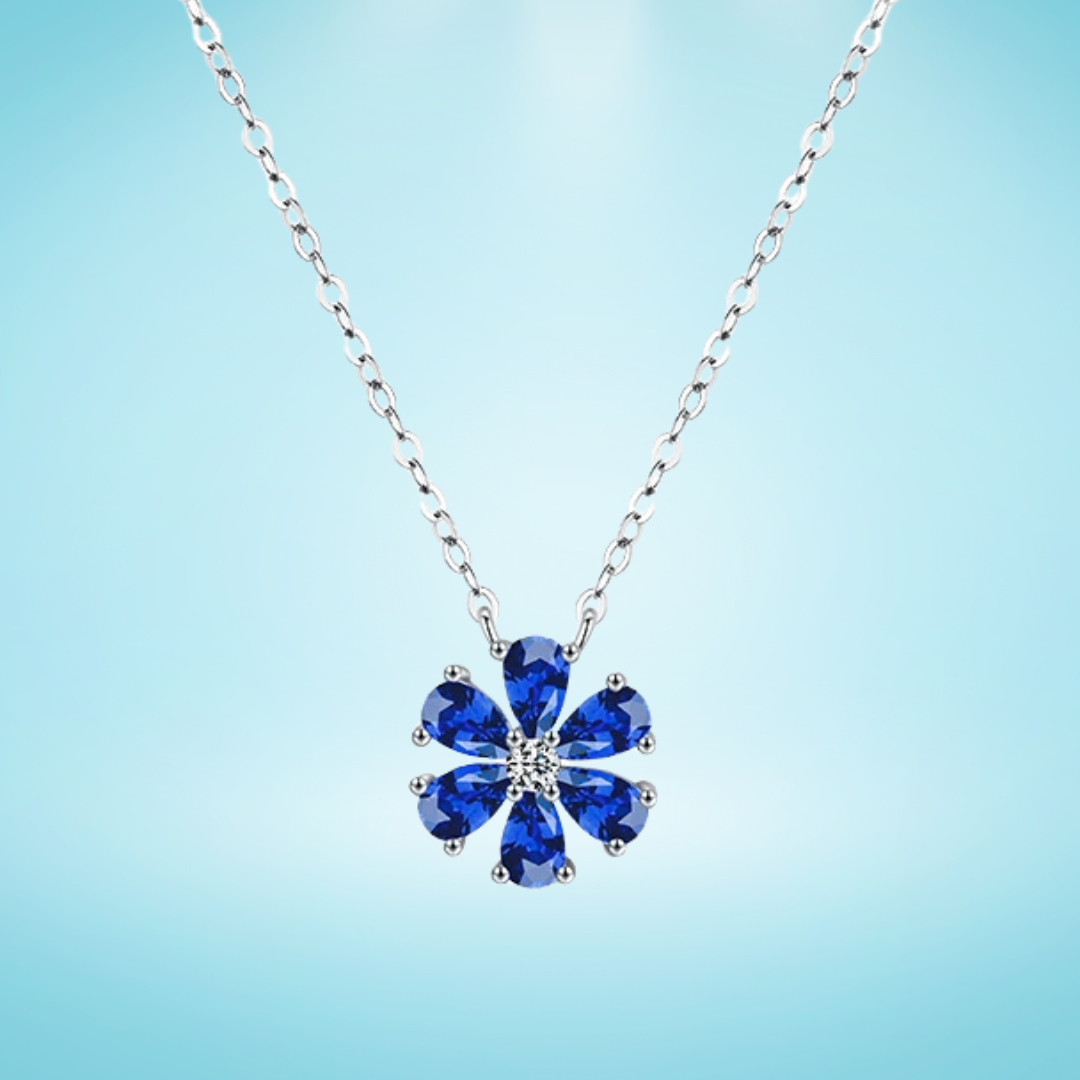 Silver necklace with a flower-shaped pendant featuring deep blue teardrop gemstones and a central white stone, on a light blue background