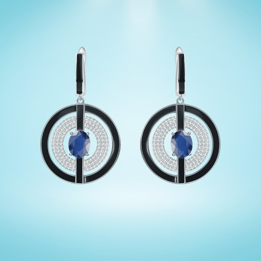Sterling silver drop earrings with black enamel circular frames, pavé-set crystal detailing, and deep blue oval gemstones at the center, on a light blue background