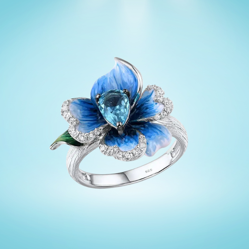 Silver ring with a sculpted blue enamel flower and a central teardrop-shaped blue gemstone surrounded by white stones

