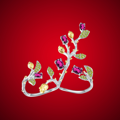 Double-finger ring with branch-like silver bands, deep pink stones, golden buds, and green pavé leaves from the Rossa Collection.

