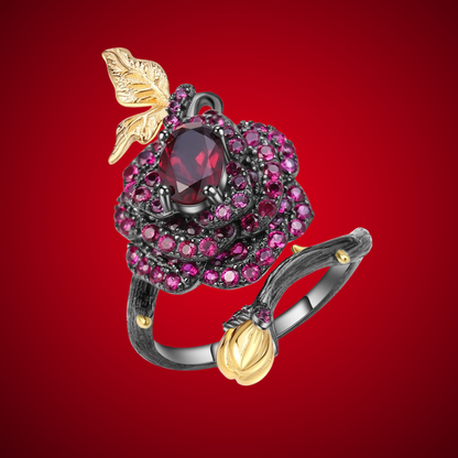 Black rose ring with layered red gemstones, accented by a gold leaf and bud on a dark branch-like band.

