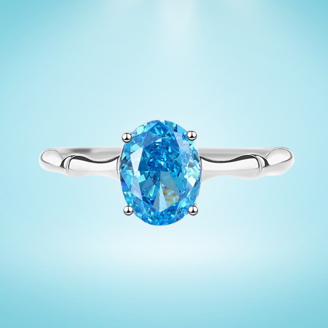 Silver ring with a vivid oval-cut blue gemstone set in a simple band, displayed on a light blue background

