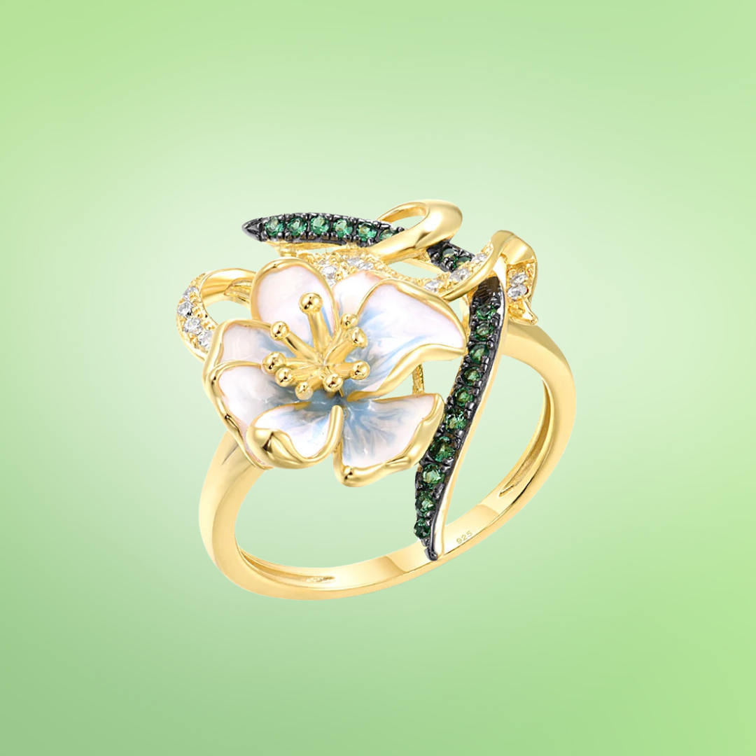 Gold ring with white flower, green crystal accents, and ribbon-like detailing

