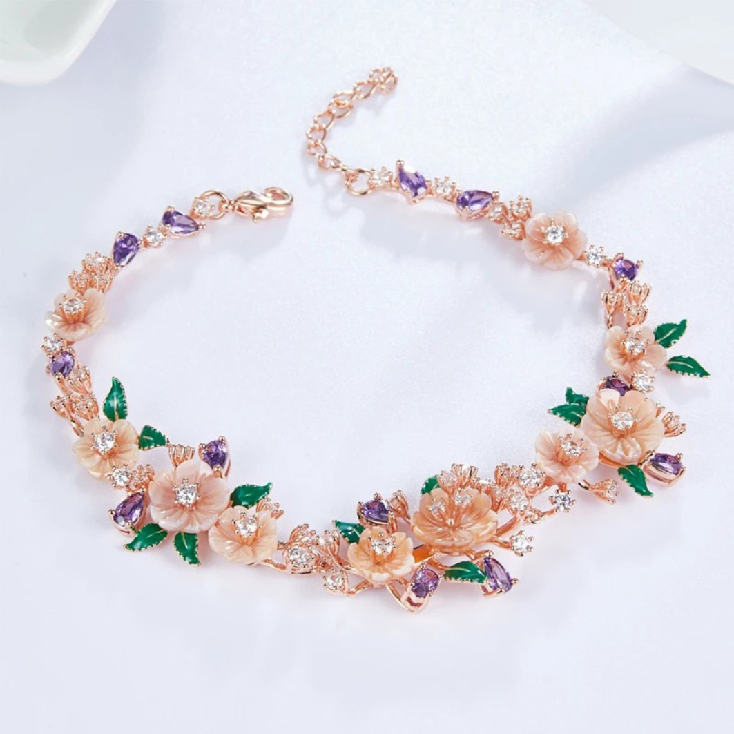 Floral bracelet with soft pink blossoms, green enamel leaves, and purple heart-shaped stones on a rose gold chain, set against a red background.


