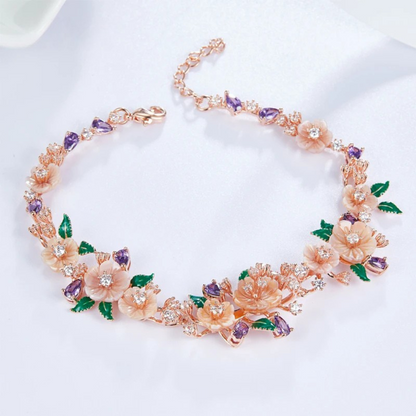 Floral bracelet with soft pink blossoms, green enamel leaves, and purple heart-shaped stones on a rose gold chain, set against a red background.

