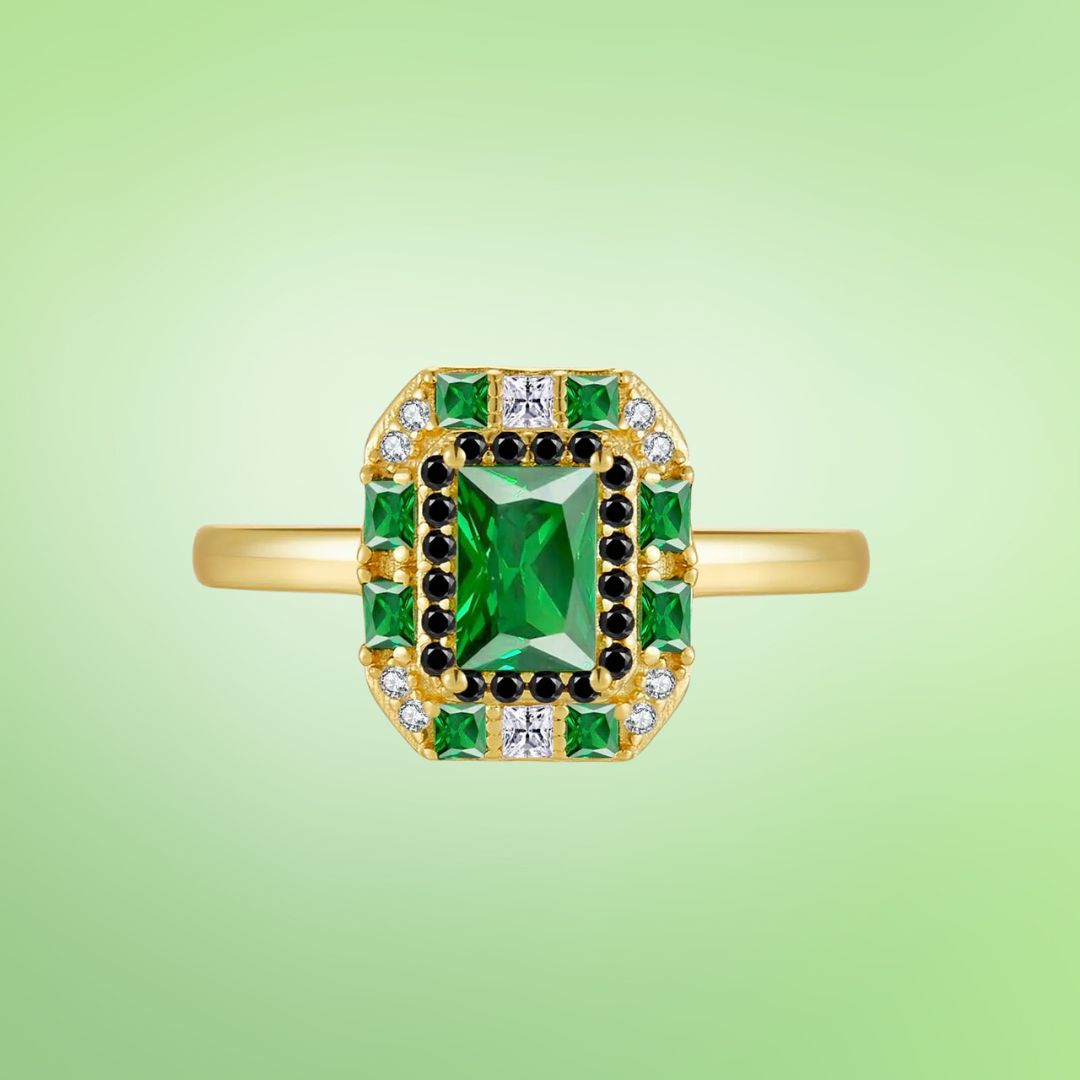 Gold ring with a central emerald-green rectangular stone, surrounded by alternating black and white crystals and square-cut green gems.

