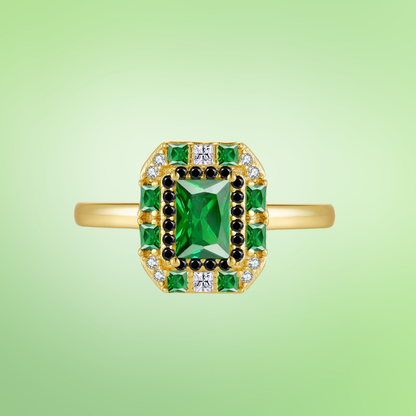 Gold ring with a central emerald-green rectangular stone, surrounded by alternating black and white crystals and square-cut green gems.

