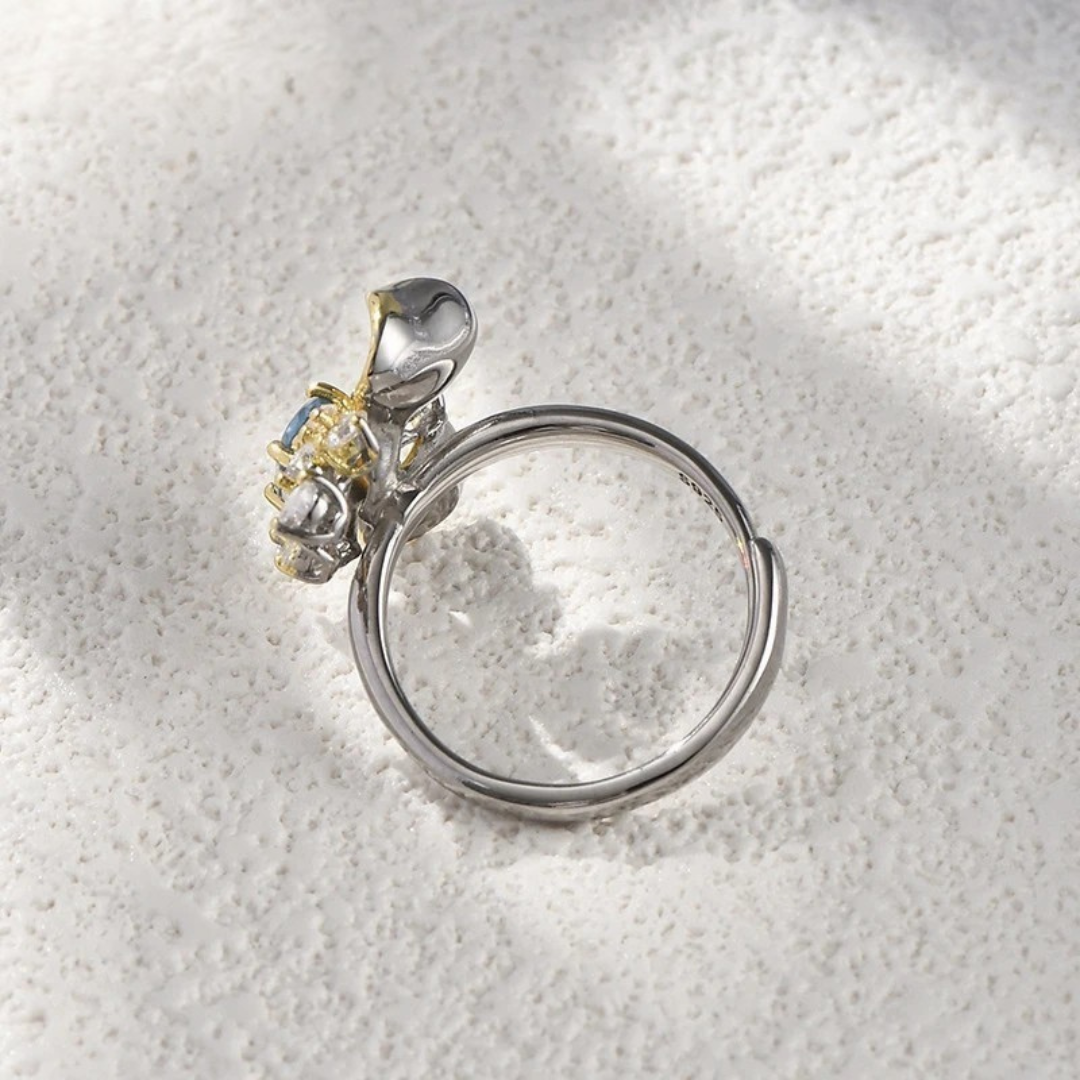 Two-tone floral ring with a blue oval gemstone, gold petal accents, and white crystal details on a silver band, set against a light blue background