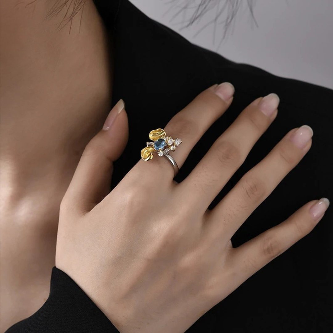 Two-tone floral ring with a blue oval gemstone, gold petal accents, and white crystal details on a silver band, set against a light blue background
