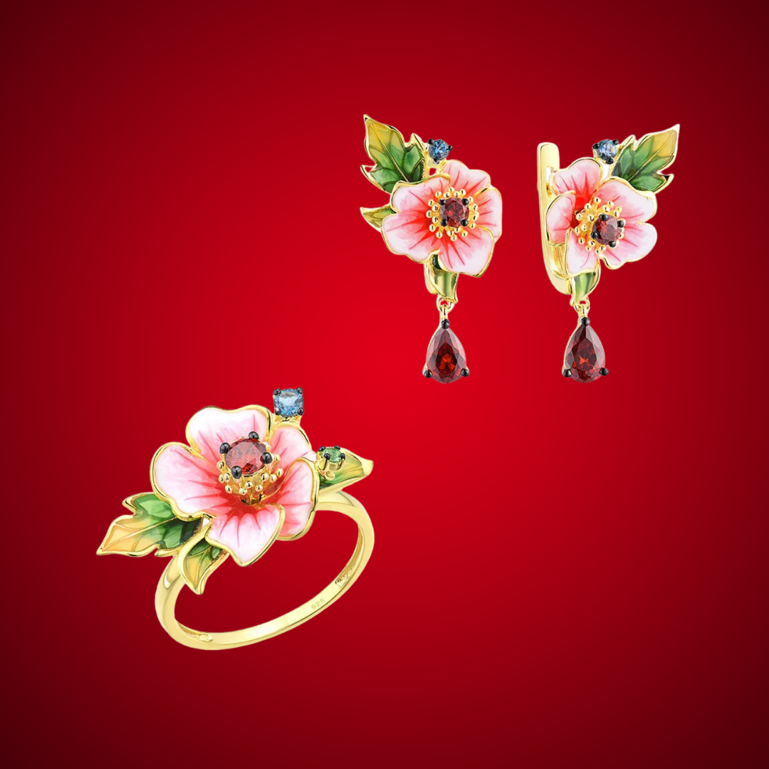 Amaryllis jewelry set with pink enamel flowers, red teardrop stones, green leaf accents, and small blue gemstones on a gold base.


