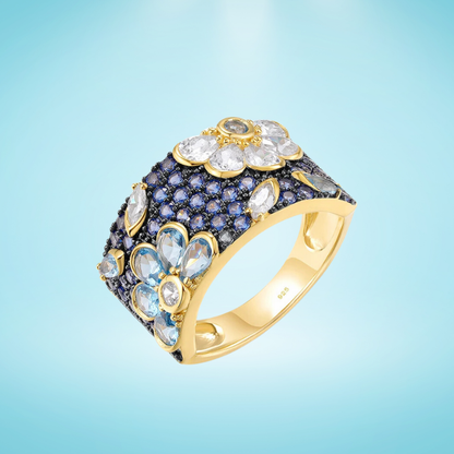 Gold-plated floral ring with a wide band, set with clear, blue, and navy gemstones forming flower shapes

