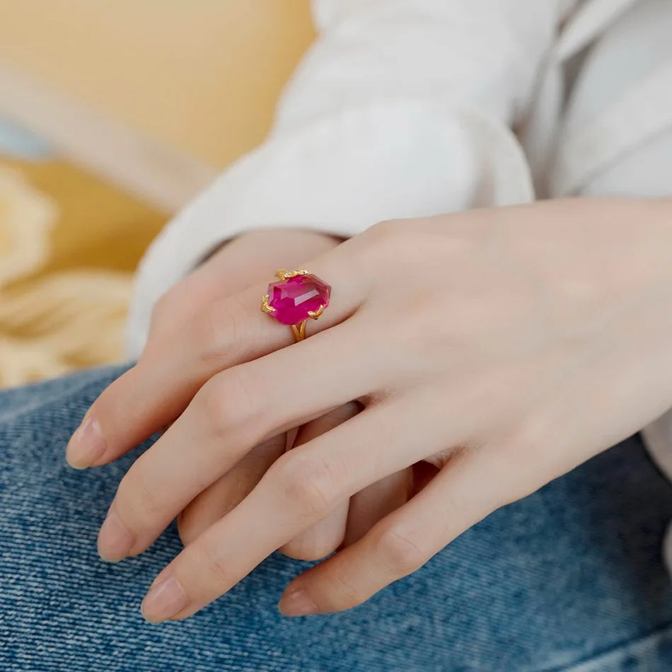 Gold ring with a bold, faceted pink gemstone held by petal-shaped prongs, displayed on a red background.

