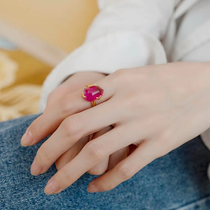 Gold ring with a bold, faceted pink gemstone held by petal-shaped prongs, displayed on a red background.

