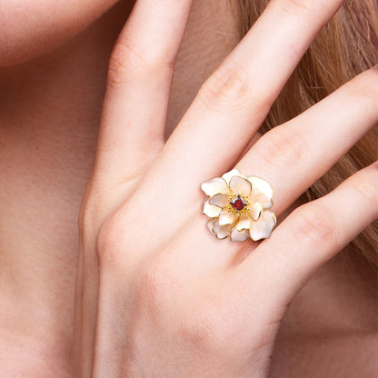 Gold ring with layered white enamel flower petals and a red gemstone at the center.

