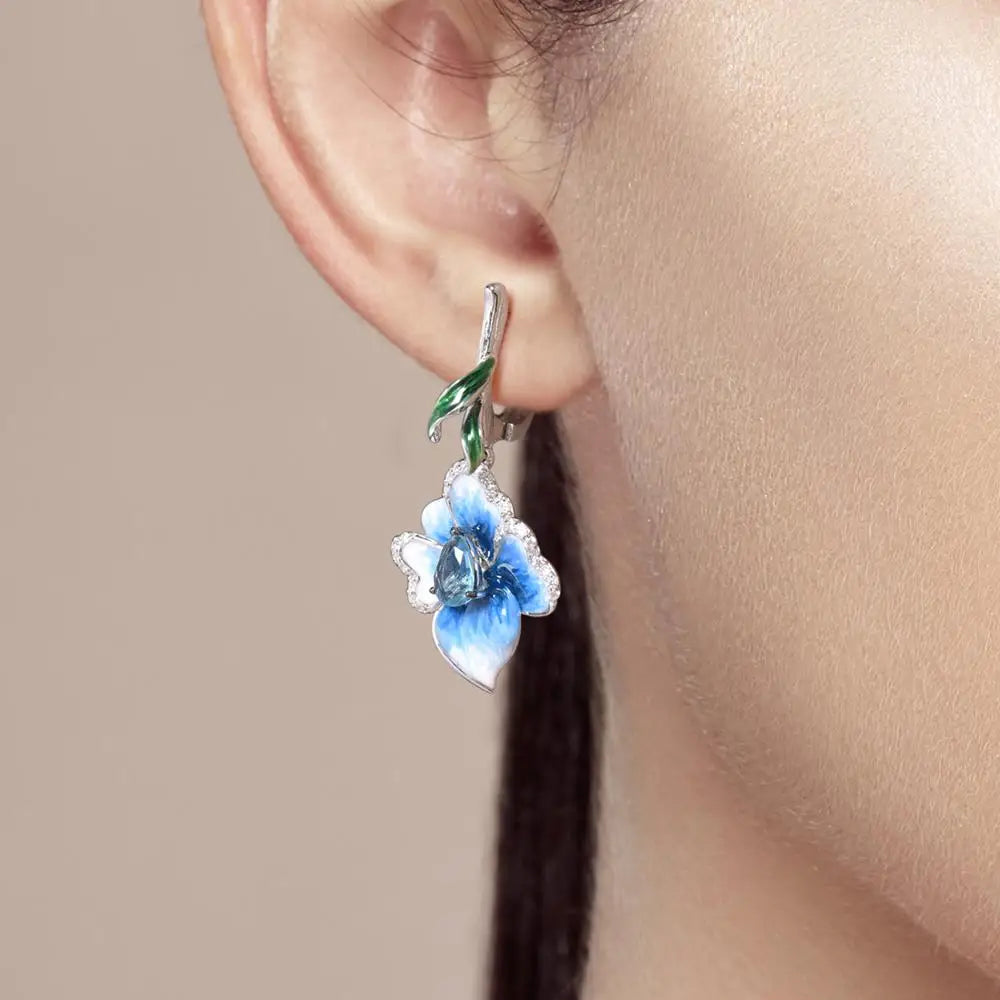 Silver floral earrings with hand-painted blue petals, green enamel accents, and teardrop blue stones.

