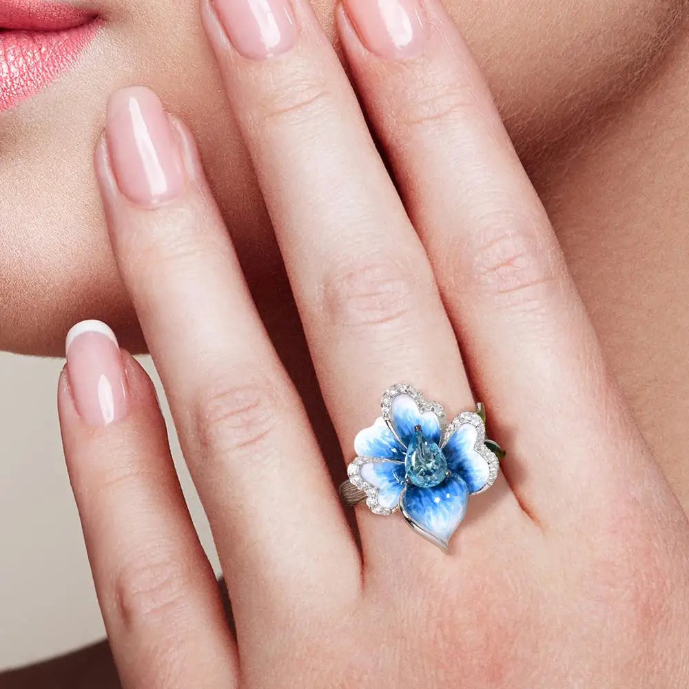 Silver ring with a sculpted blue enamel flower and a central teardrop-shaped blue gemstone surrounded by white stones

