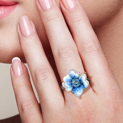 Silver ring with a sculpted blue enamel flower and a central teardrop-shaped blue gemstone surrounded by white stones


