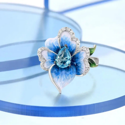 Silver ring with a sculpted blue enamel flower and a central teardrop-shaped blue gemstone surrounded by white stones

