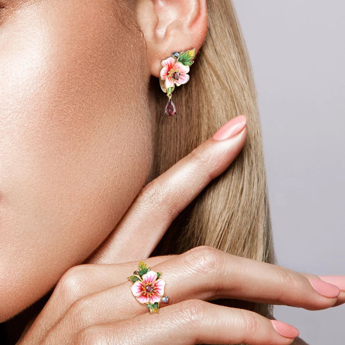 Amaryllis jewelry set with pink enamel flowers, red teardrop stones, green leaf accents, and small blue gemstones on a gold base.

