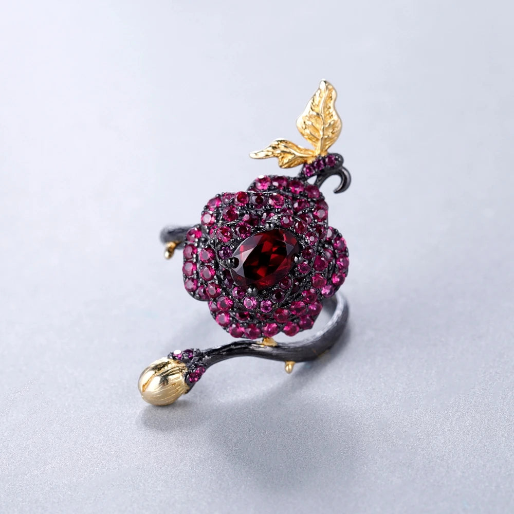 Black rose ring with layered red gemstones, accented by a gold leaf and bud on a dark branch-like band.

