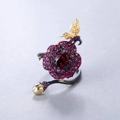 Black rose ring with layered red gemstones, accented by a gold leaf and bud on a dark branch-like band.

