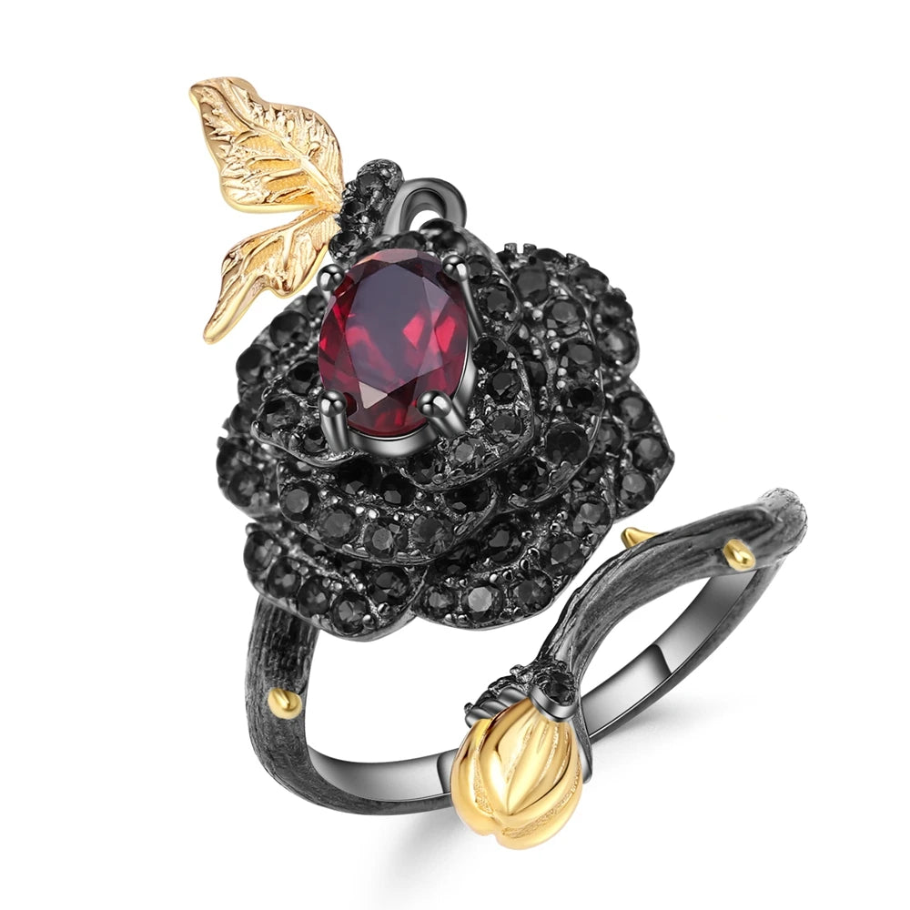 Black rose ring with layered red gemstones, accented by a gold leaf and bud on a dark branch-like band.

