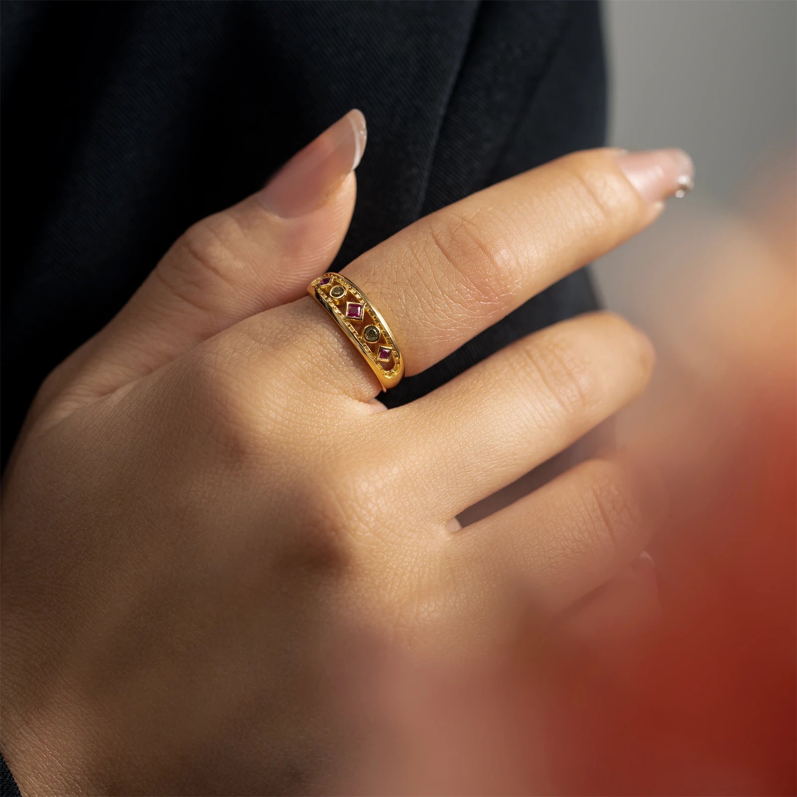 Gold ring with alternating pink square-cut and green round gemstones, framed by beaded detailing, displayed on a red background.

