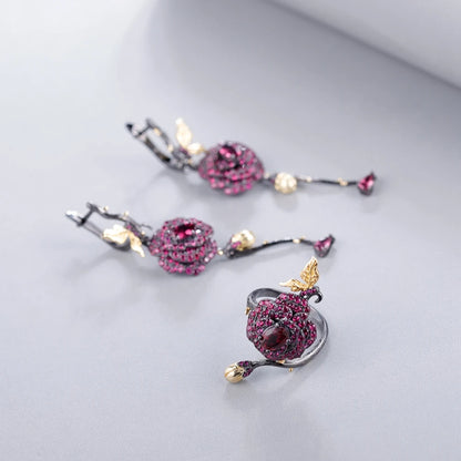 Black rose ring with layered red gemstones, accented by a gold leaf and bud on a dark branch-like band.

