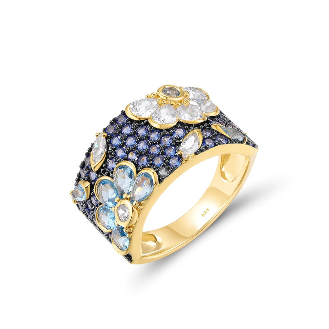 Gold-plated floral ring with a wide band, set with clear, blue, and navy gemstones forming flower shapes

